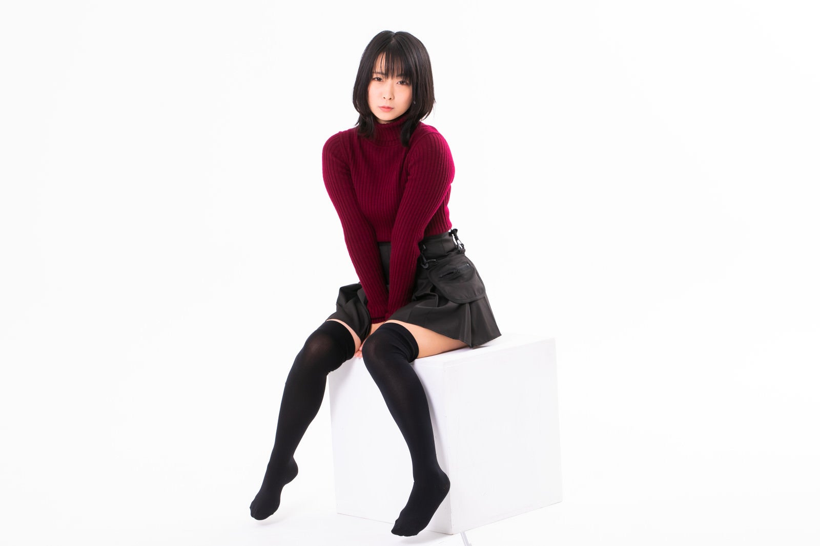 赤いニットとグレーのスカートを着た女性が内ももに手を挟んで座っている［モデル：にゃるる］