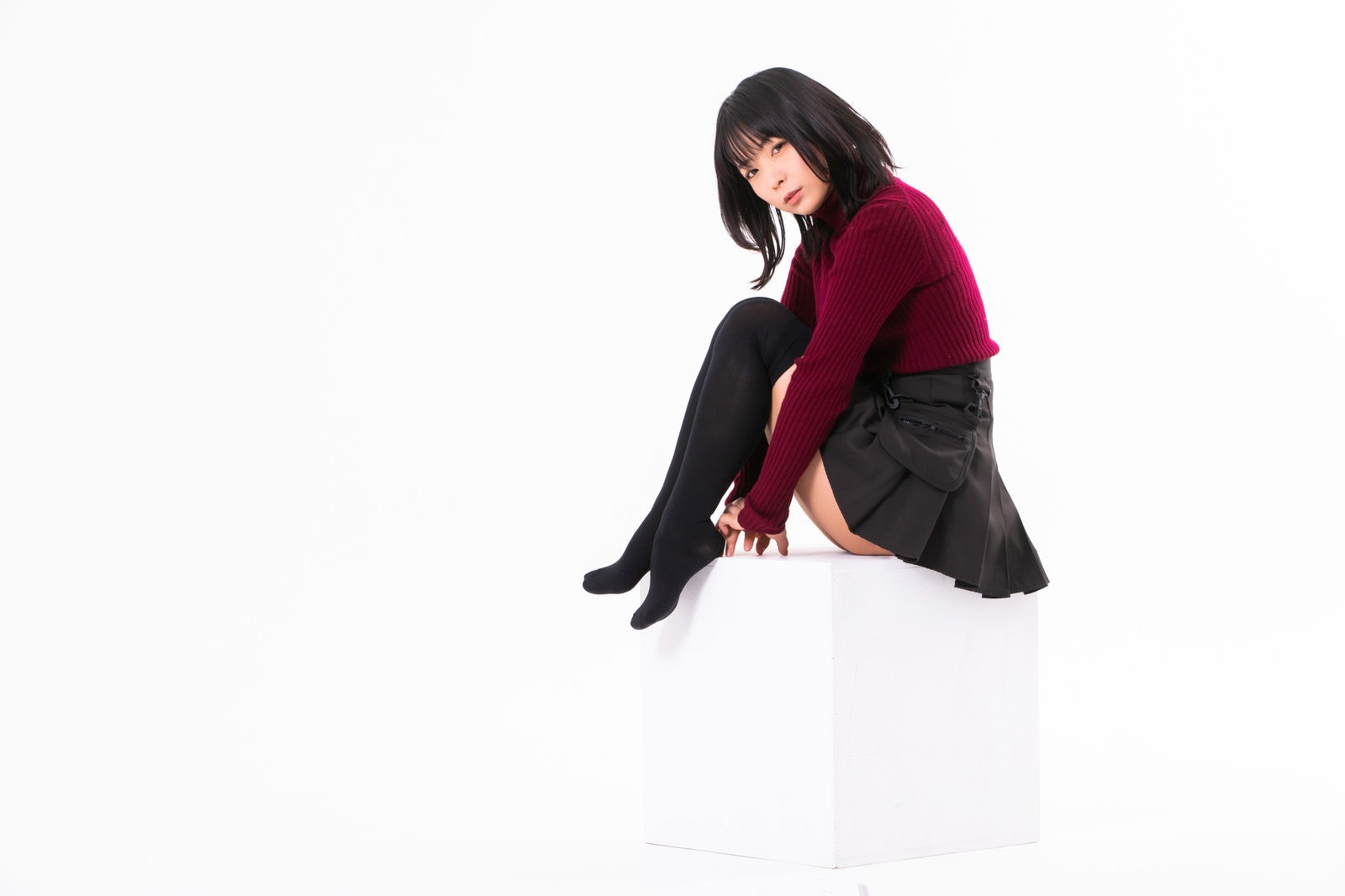 白背景のスタジオで白い箱に座りカメラを見つめるエンジ色ニットの黒髪女性［モデル：にゃるる］