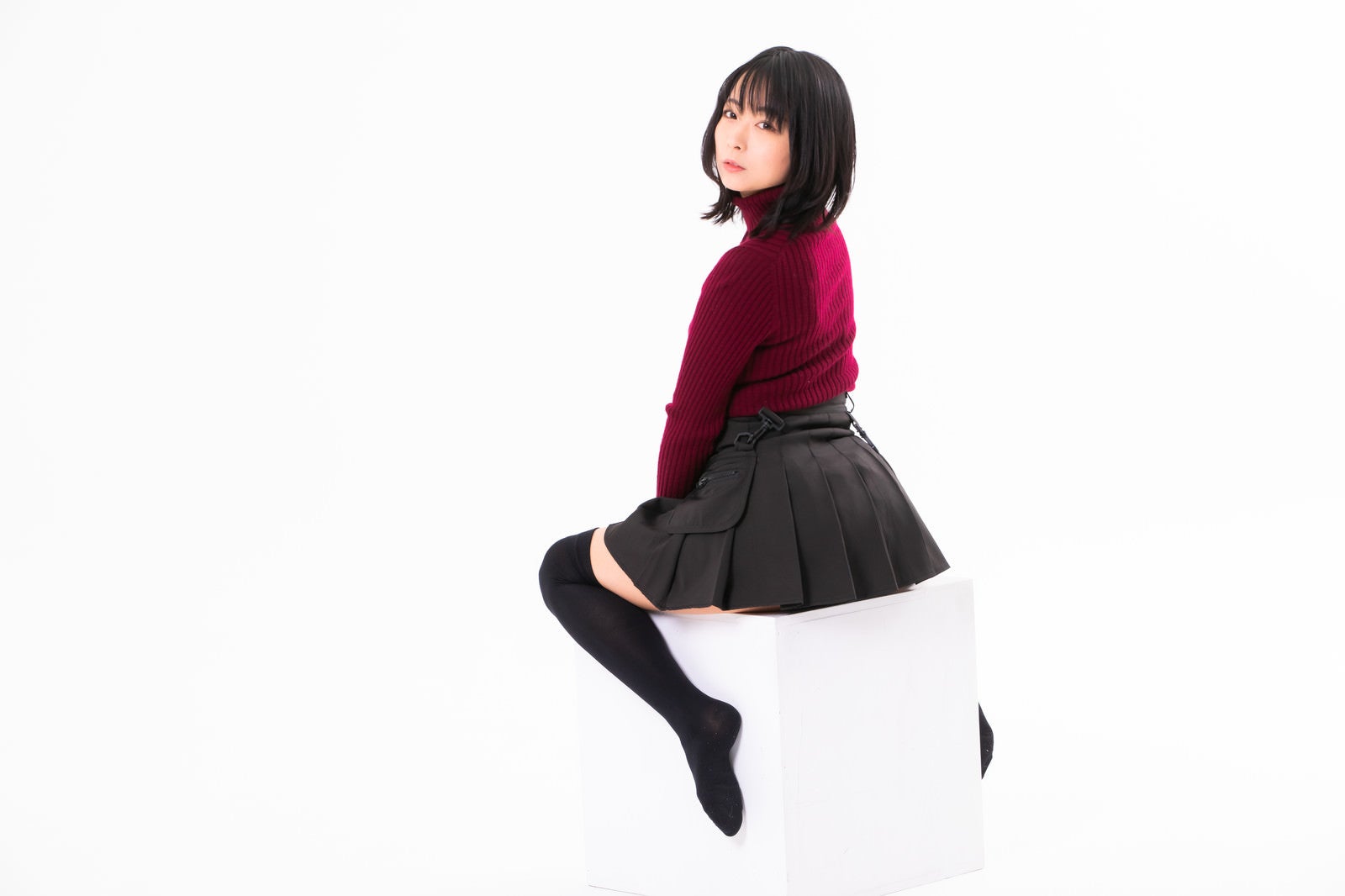 白い箱に跨って座り振り返る赤いタートルネックの女性のスタジオポートレート［モデル：にゃるる］