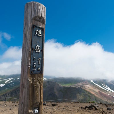 背景に雲が流れる旭岳山頂の木碑の写真