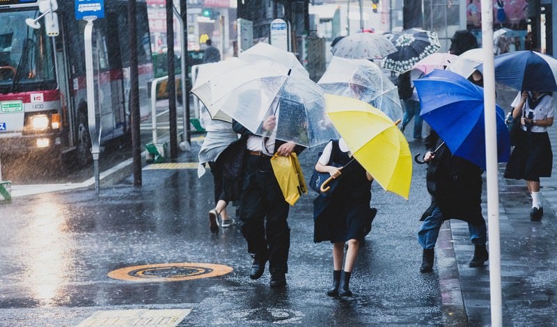 台風で突風が吹く駅前と必死に傘をさす人々の写真