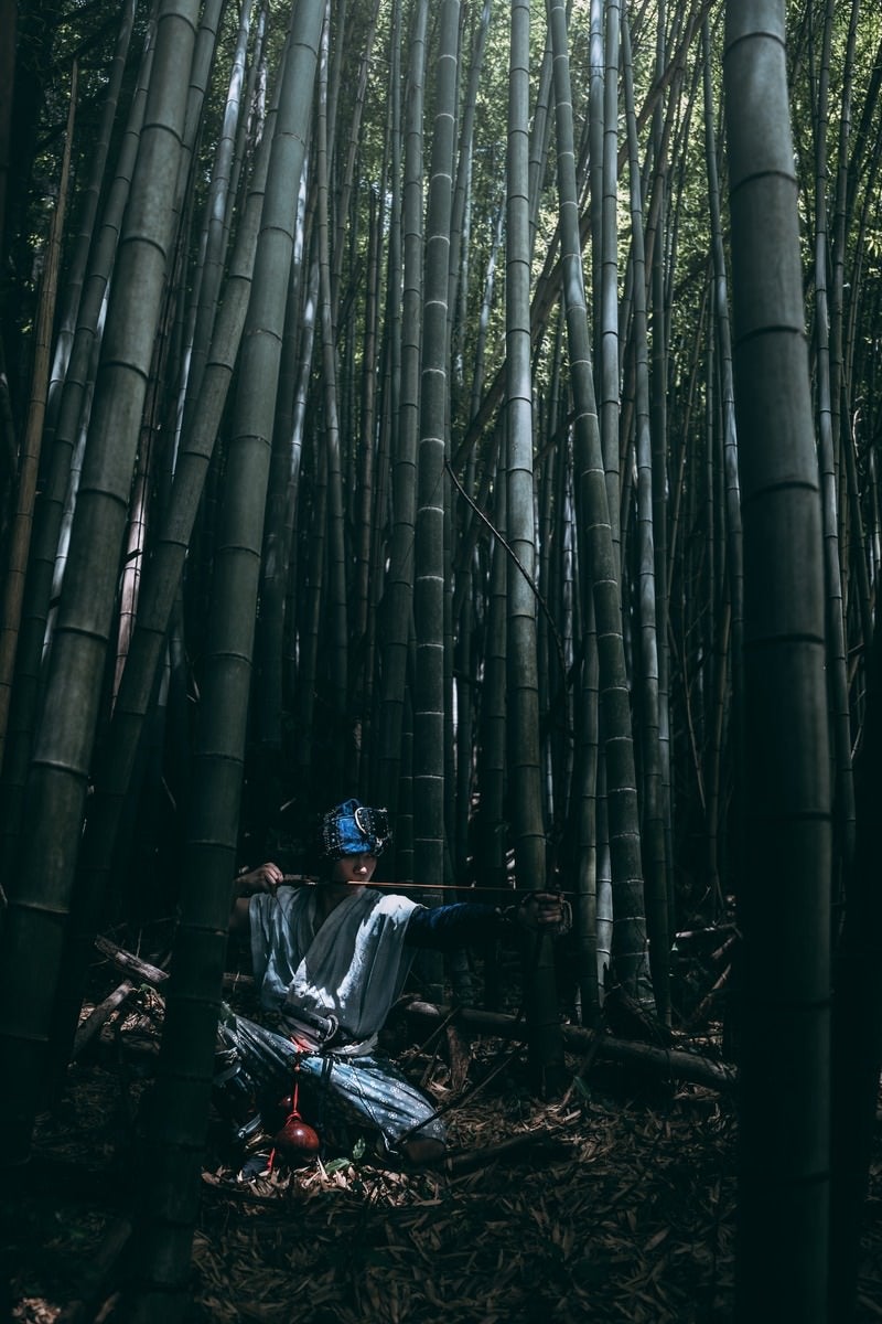 竹林の中で木漏れ日を浴びながら大弓を引く野盗の男性［モデル：まーこ］