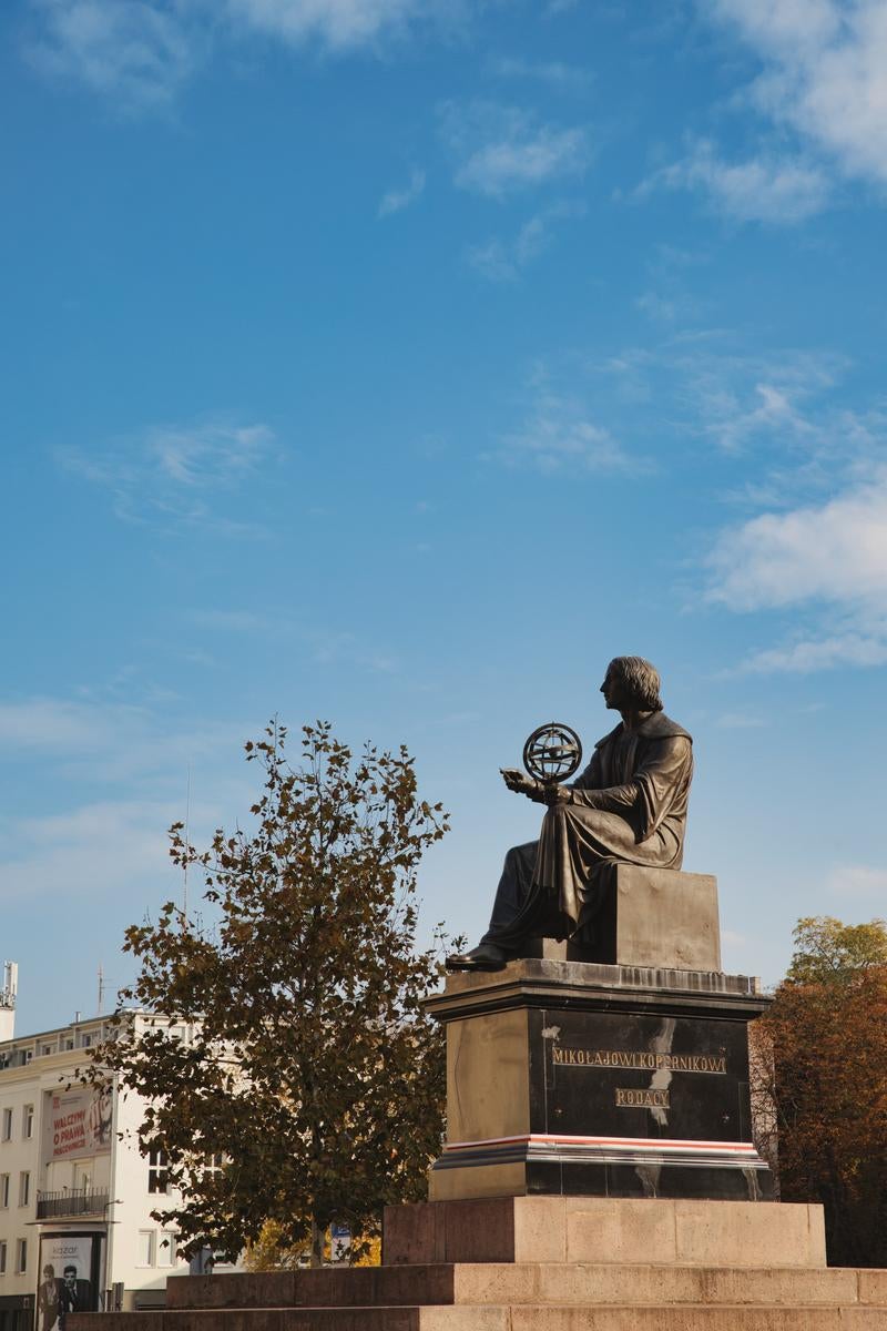 ワルシャワの広場に立つコペルニクス像。銅製の像が球体を持ち、背後に青空と秋の樹木が見える