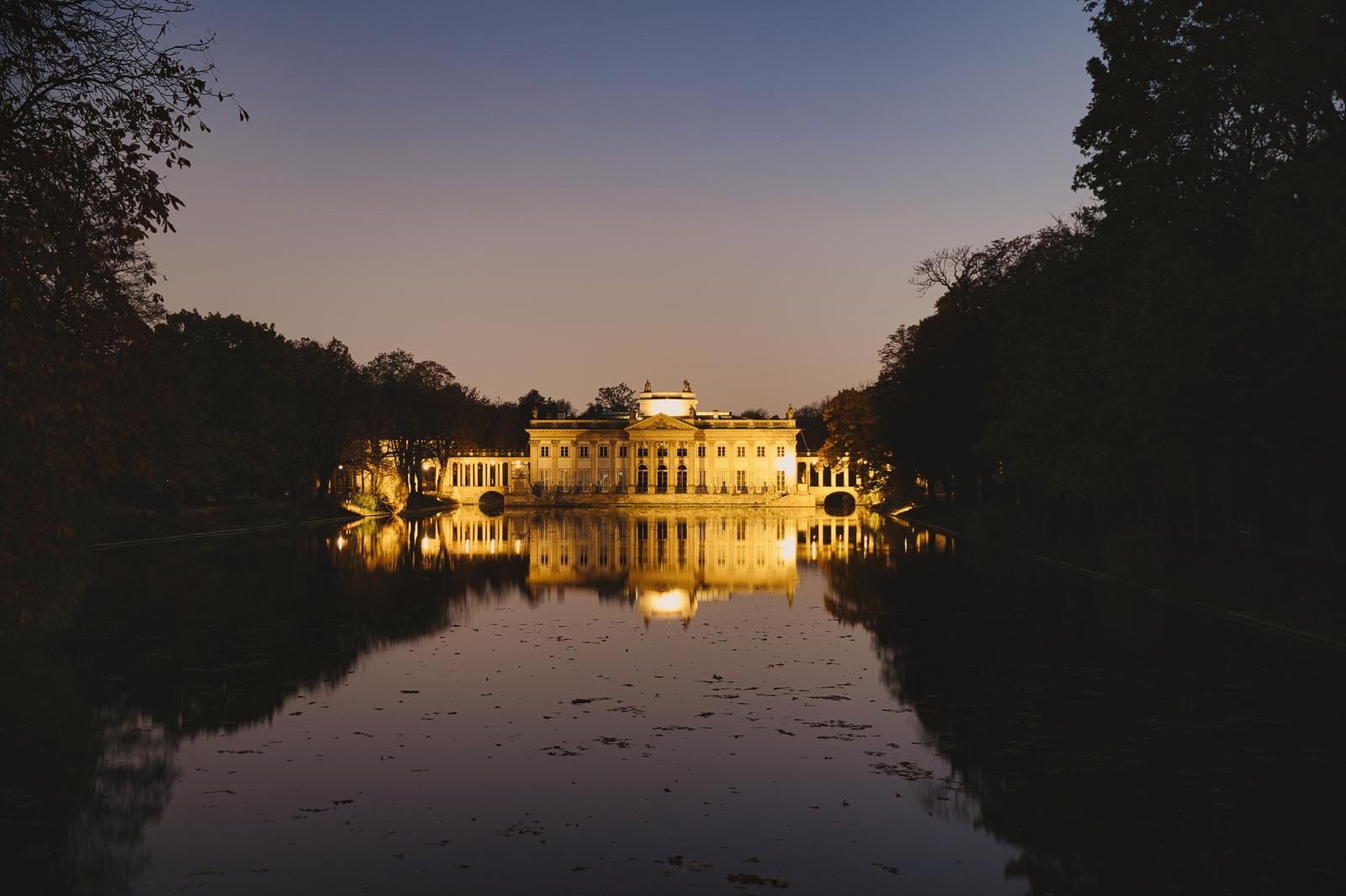 夕暮れ時にライトアップされたワルシャワのワジェンキ宮殿が池の水面に映り込んでいる景観