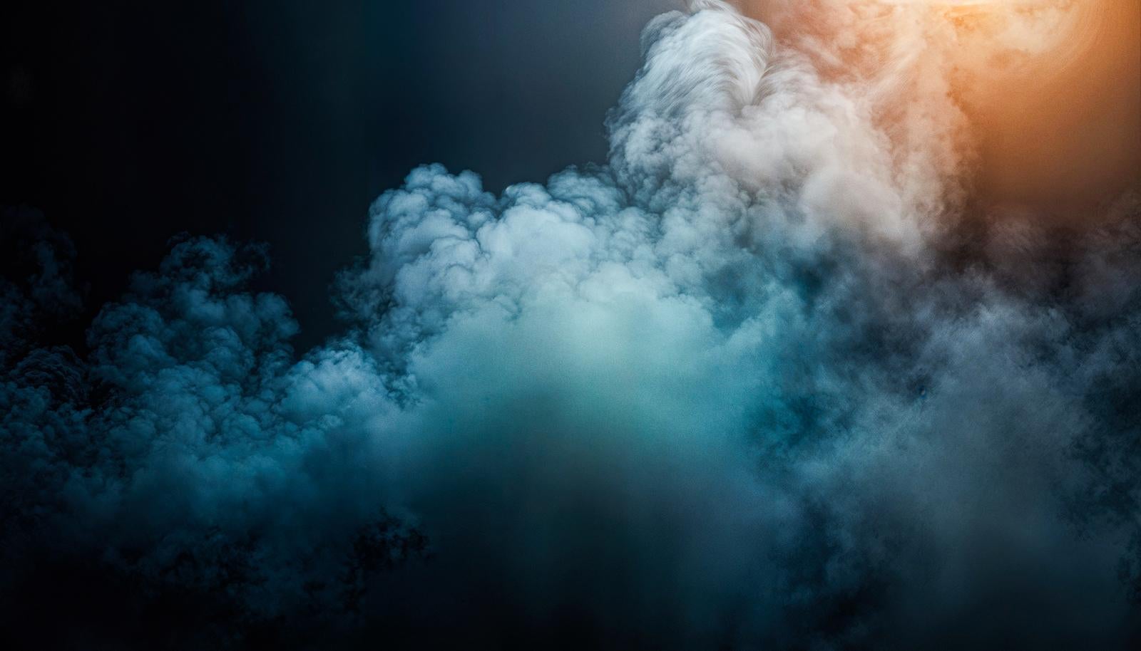 暗い背景の中で右上からオレンジの光を受けながら大きく広がる青みがかった煙