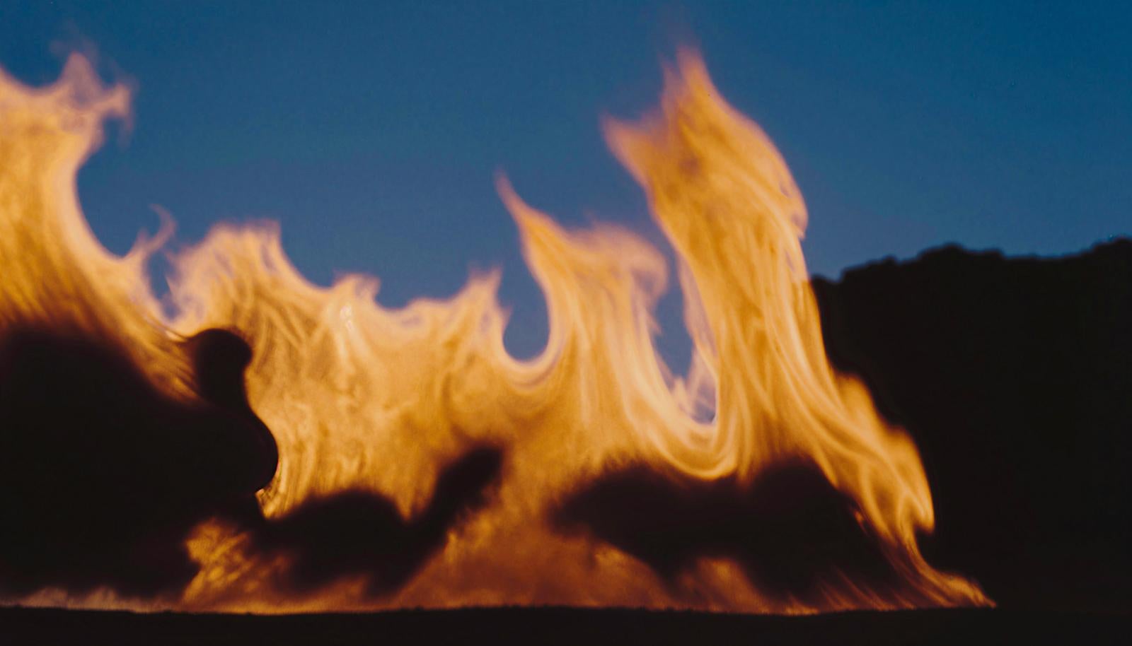 Campfire (takibi) with yellow and orange flames rising in wave-like patterns at night, with dark mountains and deep blue sky in the background
