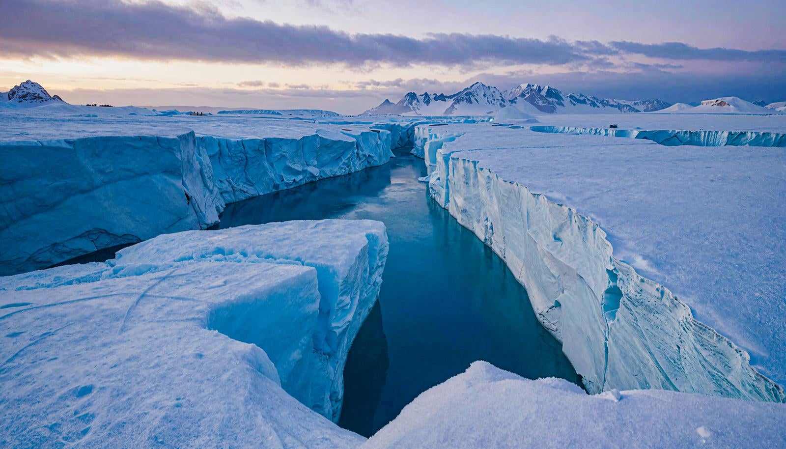 Deep blue water pooling in large glacier crevasses with snowy mountains stretching across the horizon under the twilight sky of an icy plain