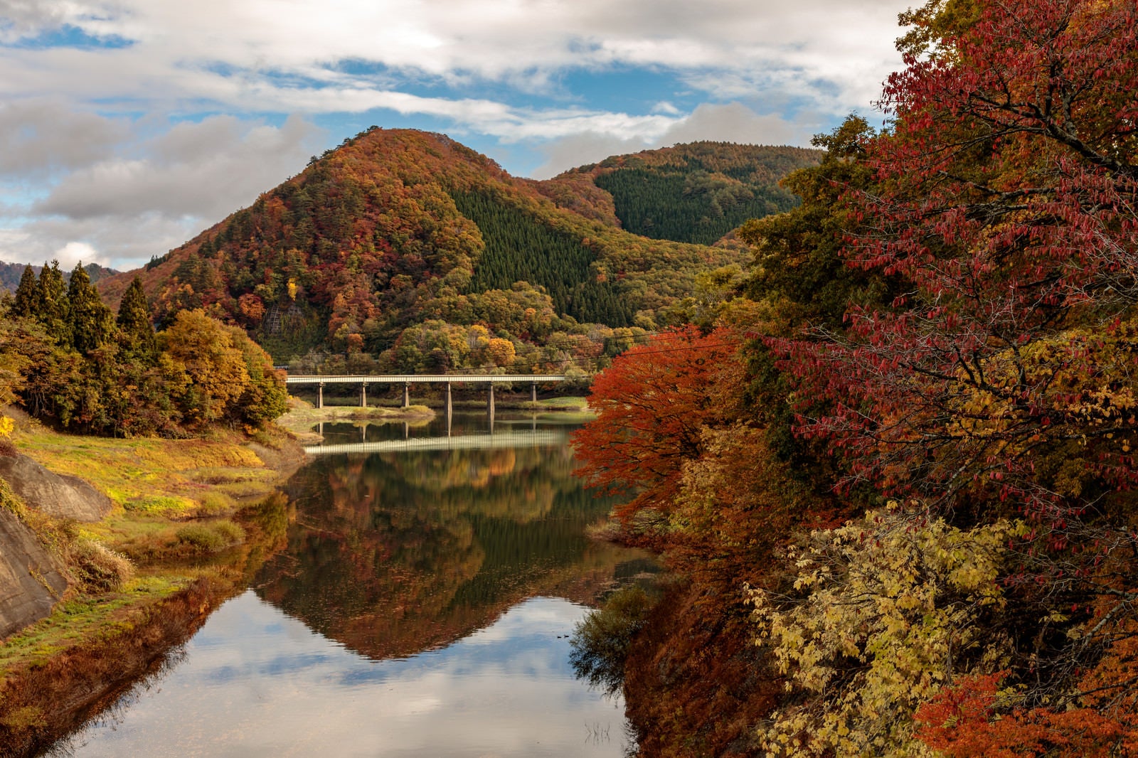 「紅葉した木々とダム湖に反射する山々と鉄橋」の写真