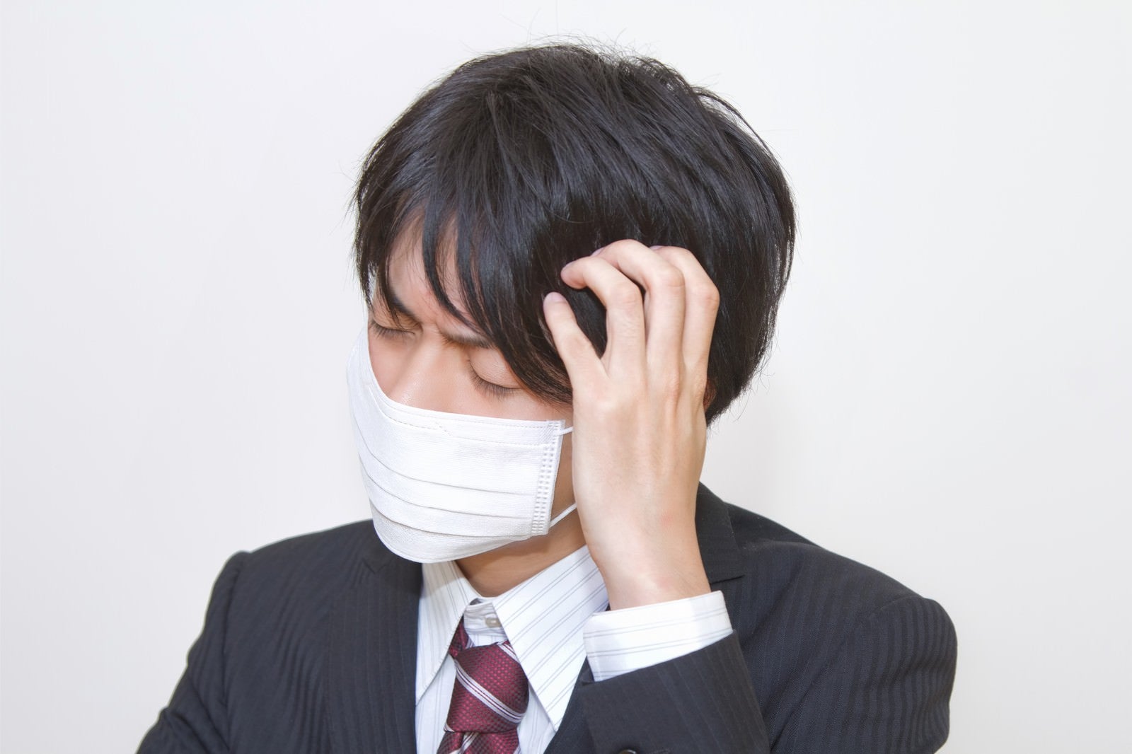 「マスクをして頭をかかえるスーツの男性」の写真［モデル：Tsuyoshi.］