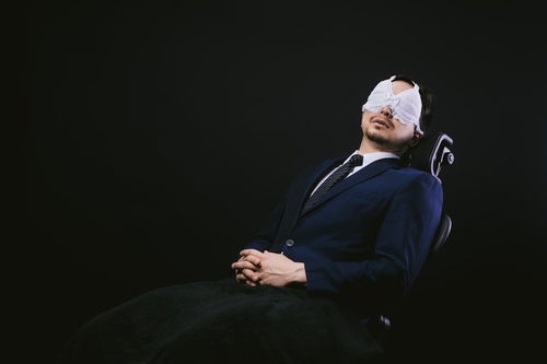 業務中にレースのアイマスクを装着して仮眠を取るサラリーマンの写真
