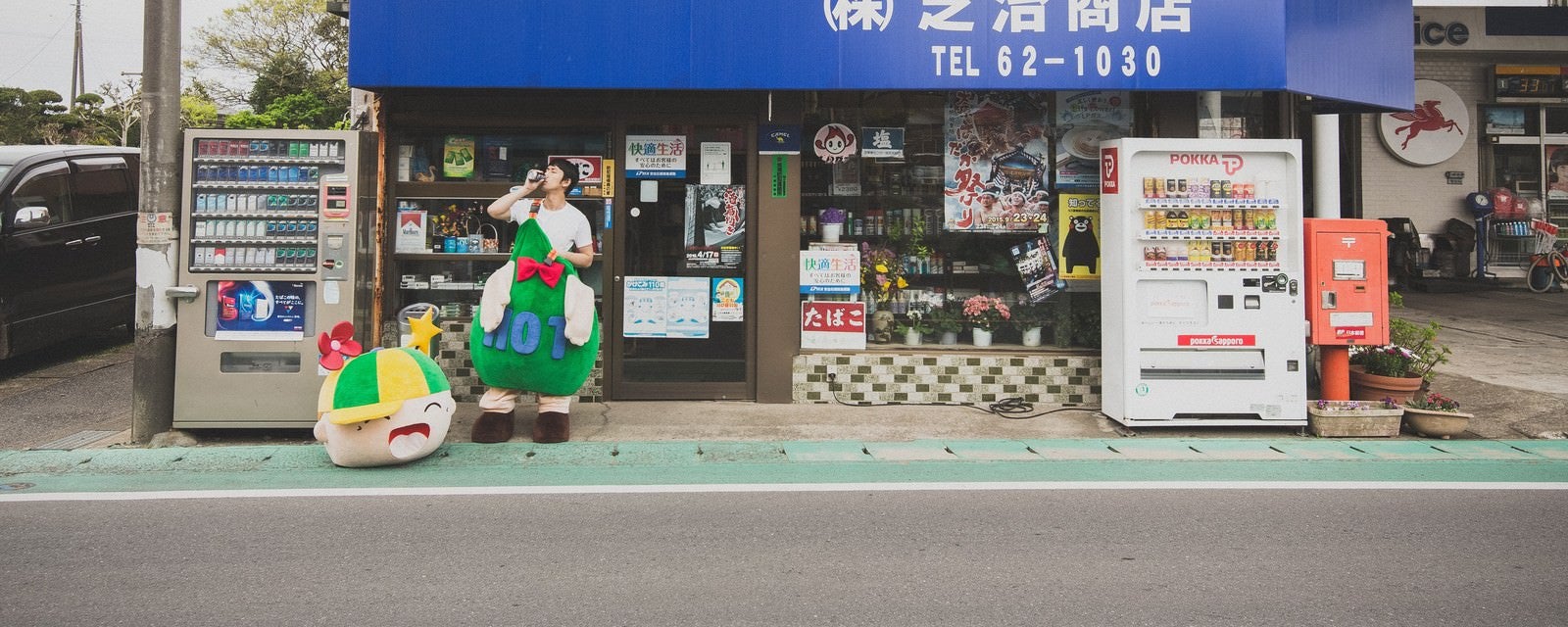 「商店の前でコーヒーを飲むゆるキャラと中の人」の写真［モデル：大川竜弥 ホットくん］