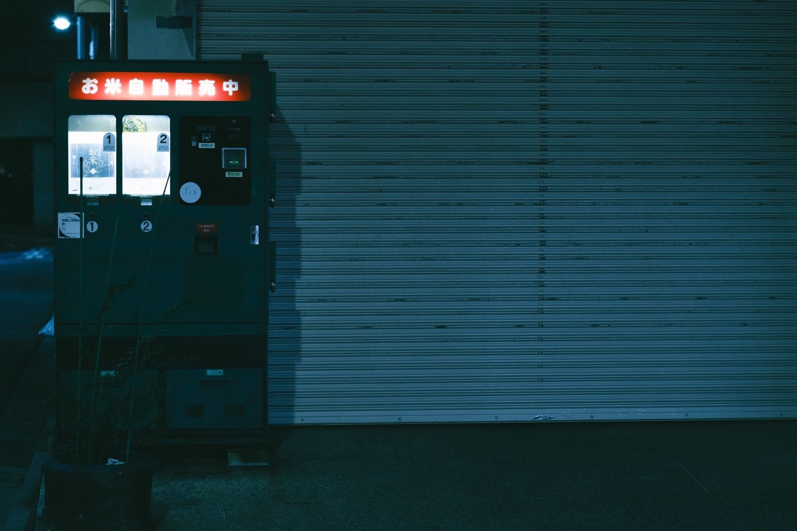 「恍惚と明かりがつく閉店後のシャッター横のお米自動販売機」の写真