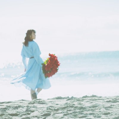 赤い花束を持ち砂浜を歩く女性の後ろ姿の写真