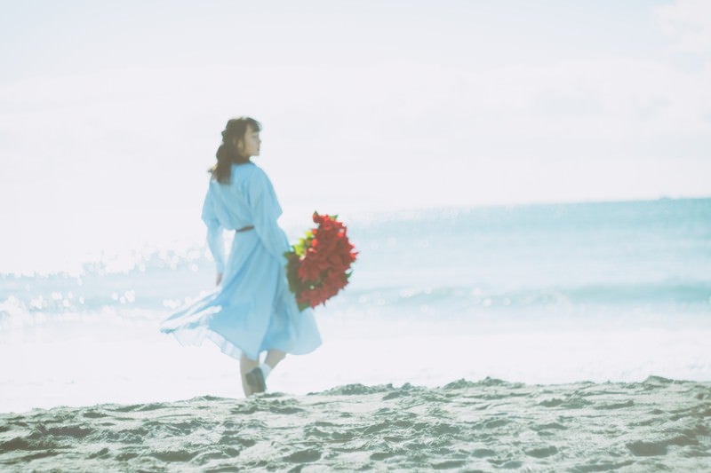 赤い花束を持ち砂浜を歩く女性の後ろ姿の写真