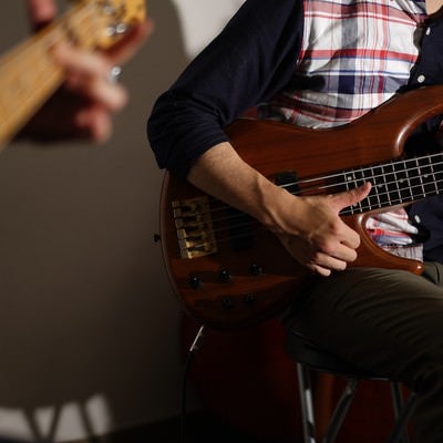 カーディガンとチェックシャツ姿の男性がベースギターを演奏する音楽活動の様子の写真