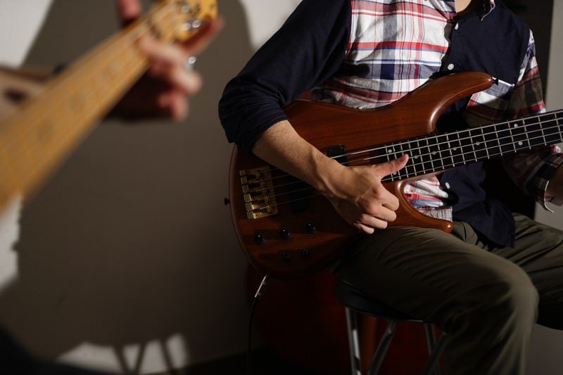 カーディガンとチェックシャツ姿の男性がベースギターを演奏する音楽活動の様子の写真