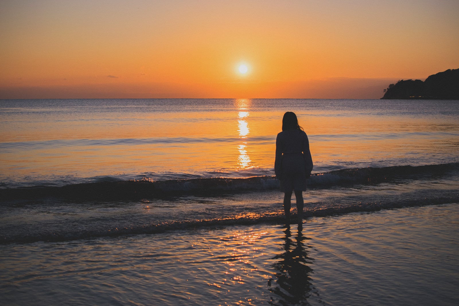 「夕日に向かって浜辺に立ちすくむ女性」の写真