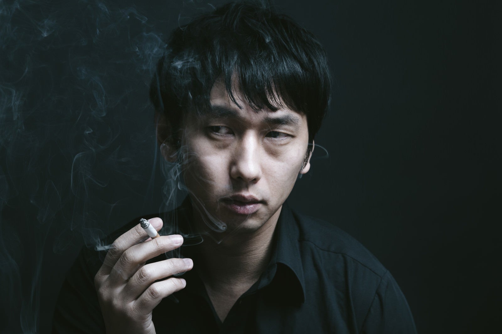 「暗い喫煙室で煙草を吸う男性」の写真［モデル：大川竜弥］