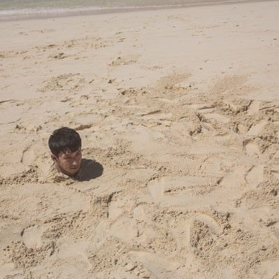 砂浜に埋まり自身の影で方角を確認する男性の写真
