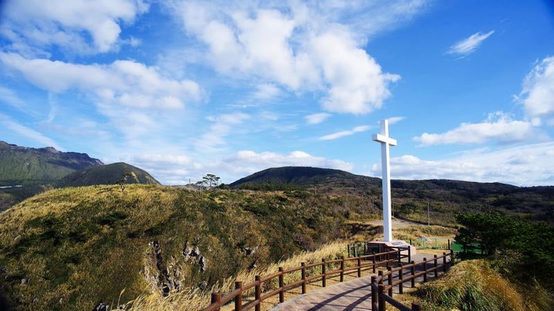 丘の上に立つ白い十字架と自然豊かな山々の写真