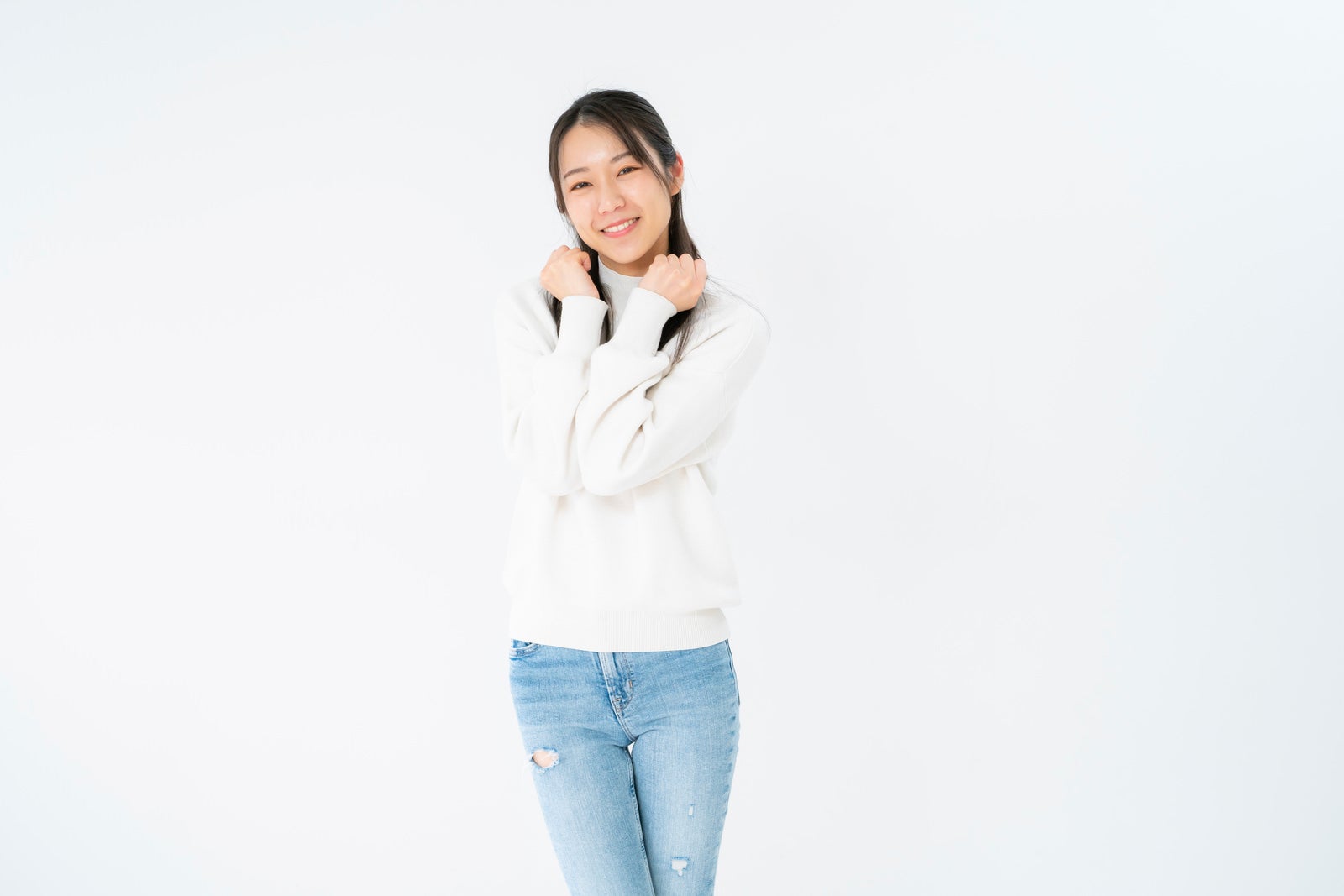 白背景の前で両手をグーにしてぶりっ子ポーズをとり笑顔を見せる白いスウェットの女性［モデル：SAKI］