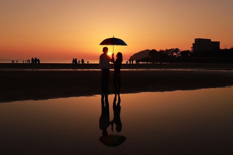 夕暮れ時に浜辺で相合傘するカップルの写真