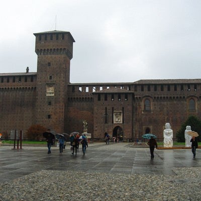 スフォルツェスコ城前の広場を行き交う傘を差す観光客（イタリア ミラノ）の写真