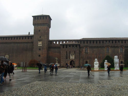スフォルツェスコ城前の広場を行き交う傘を差す観光客（イタリア ミラノ）の写真
