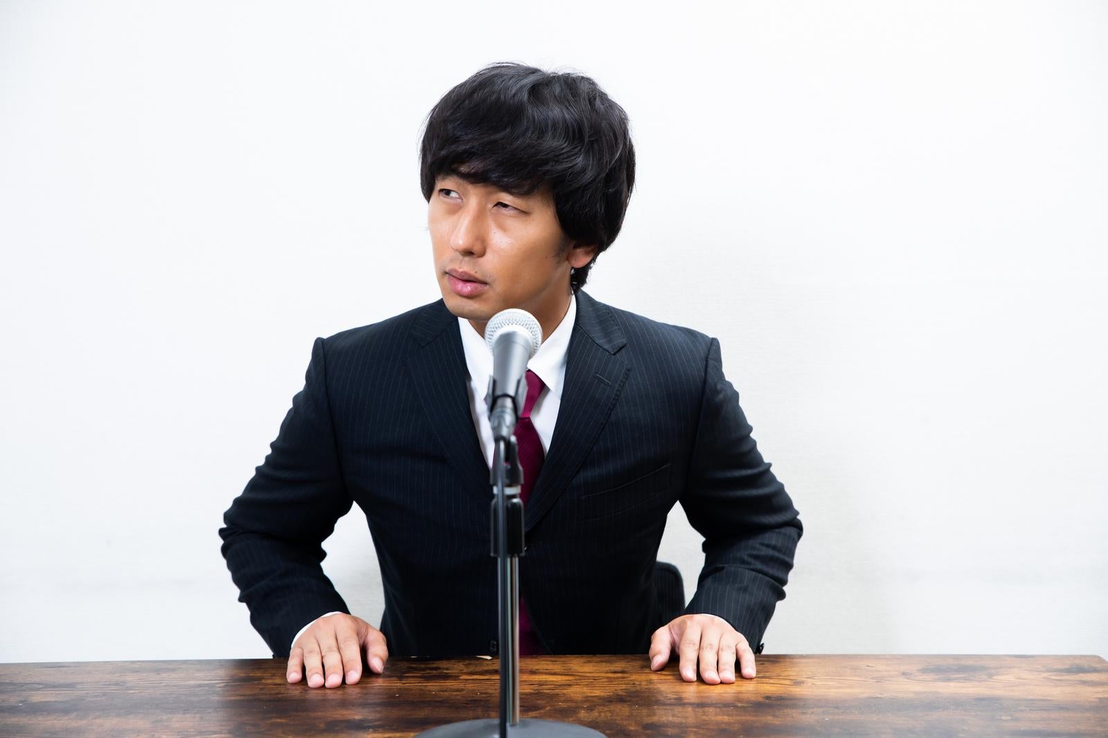 記者会見の机に座りマイクの前で横を向くスーツ姿の男性［モデル：大川竜弥］