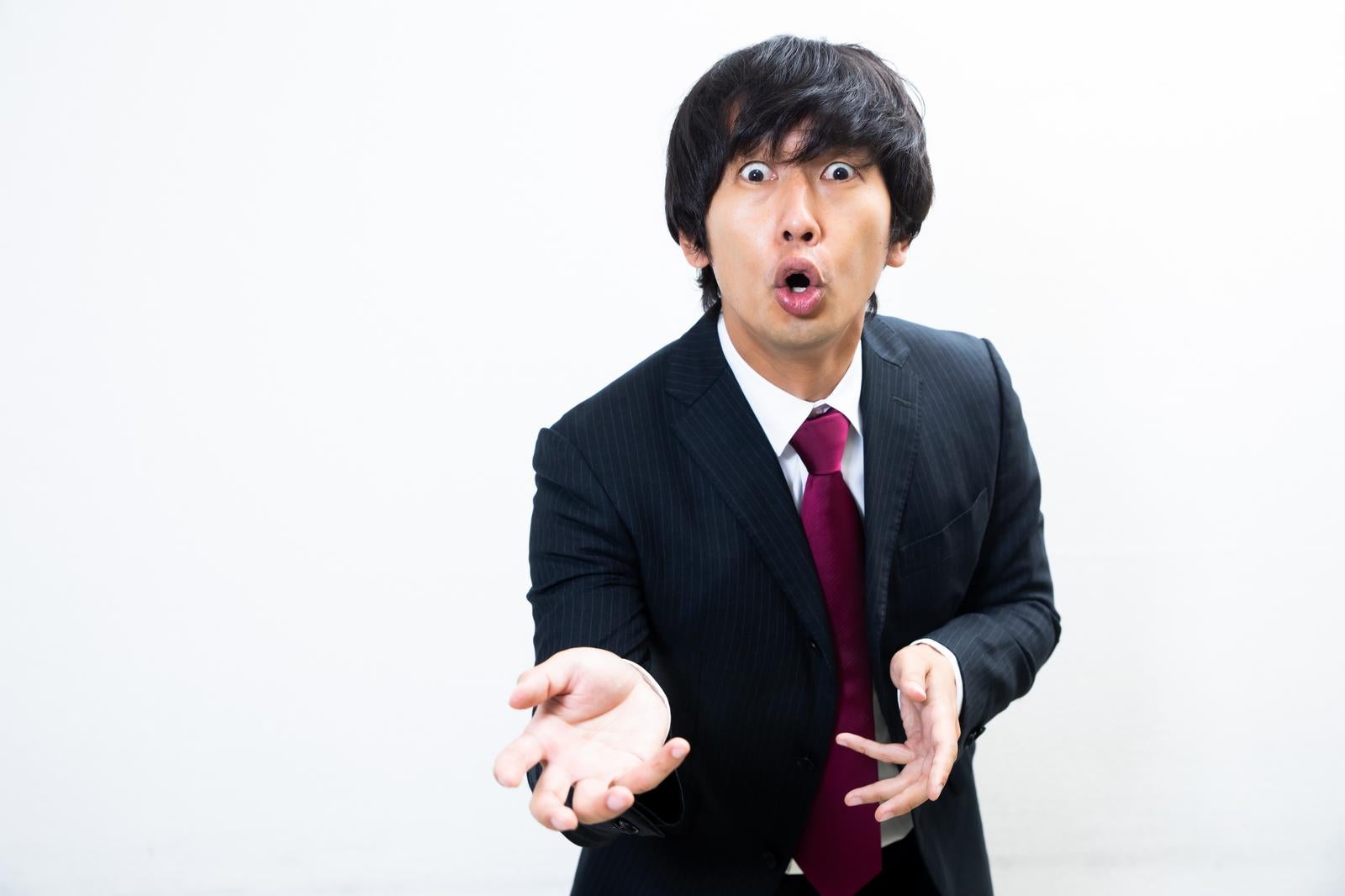 白い背景の前で両手を広げながら熱弁をふるうスーツ姿の男性［モデル：大川竜弥］