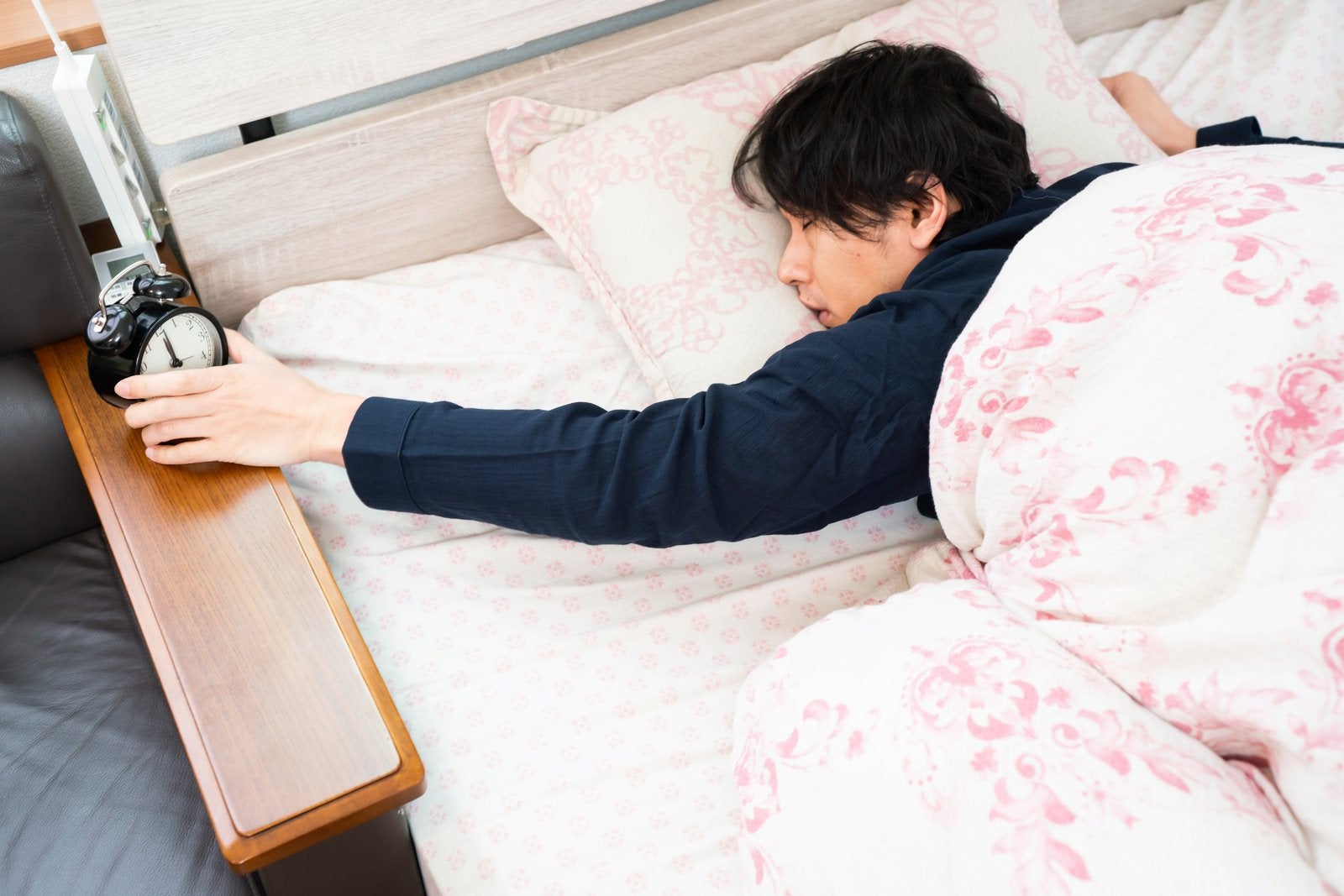 「寝ながら時計を止める寝坊男子」の写真［モデル：大川竜弥］