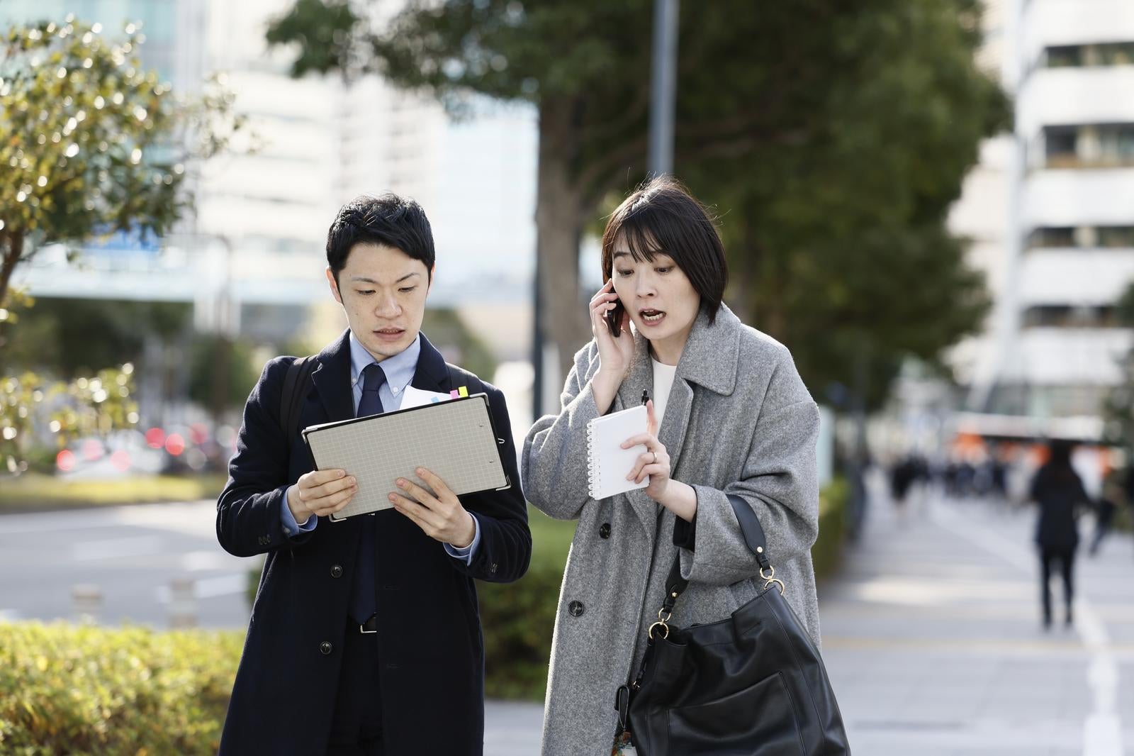 街中で電話をする女性と資料を確認する男性のビジネスパーソン［モデル：たけべともこ モデルリリース］