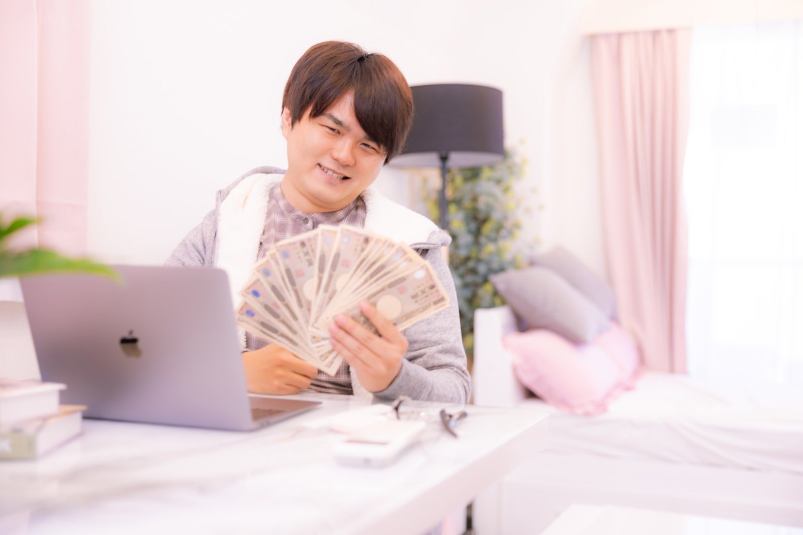現金の札束を手にして笑う男の子。背景にパソコンと観葉植物のある室内［モデル：はしも。］