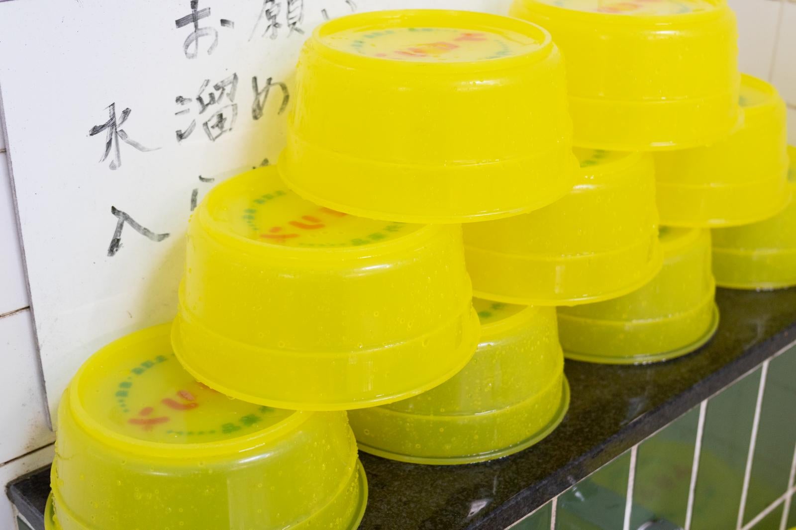 銭湯の白タイル壁の前に積み重ねられた黄色いプラスチック製の湯桶