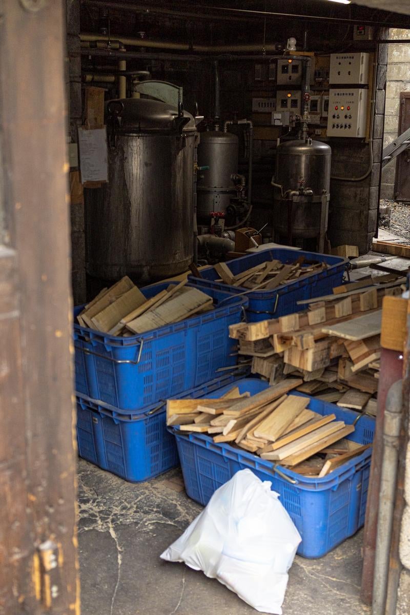 青いコンテナに積まれた薪と、奥に並ぶ黒い給湯タンクや制御盤が見える銭湯のボイラー室