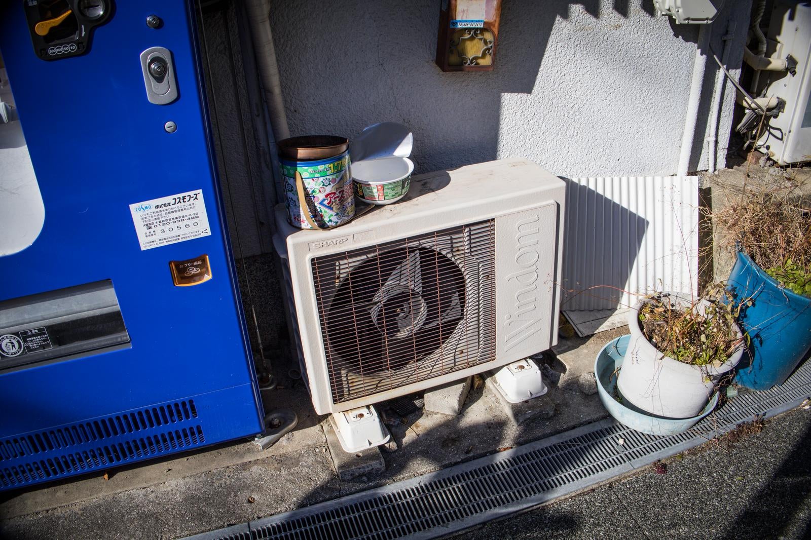 自販機の横に置かれた古いSHARP Windomの室外機と、その上に載った殺虫剤の缶とボウル