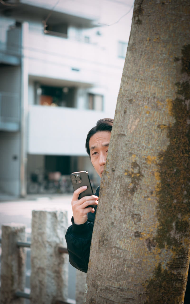「木陰に隠れながらスマホで盗撮する男性（41歳）」の写真［モデル：大川竜弥］