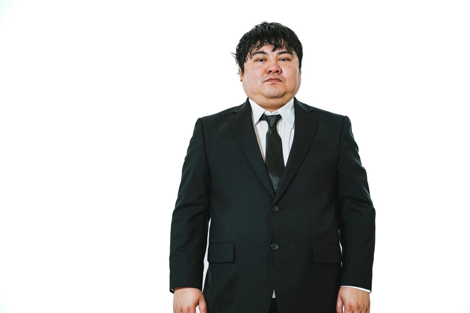 黒いストライプスーツと白いシャツ、黒いネクタイを着た男性が白背景の前で正面を向いて立っている［モデル：段田隼人］