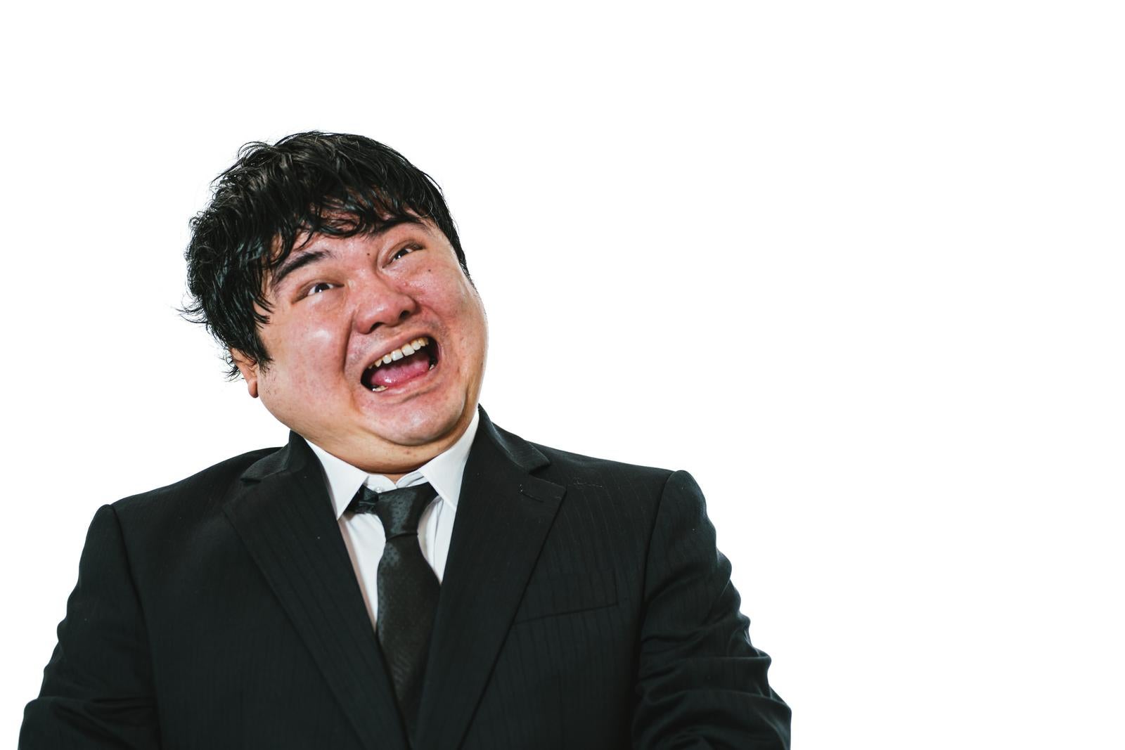 A man in a suit with disheveled hair laughing with a strained expression and wide-open mouth against a white background
