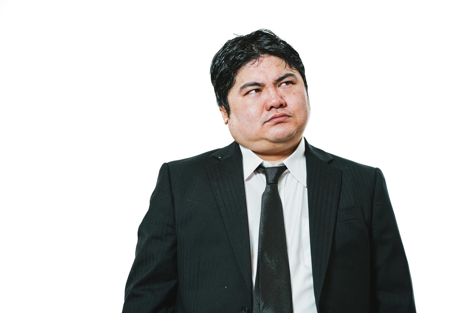 白背景の前で視線を右上に逸らしているスーツ姿の太めの男性会社員［モデル：段田隼人］