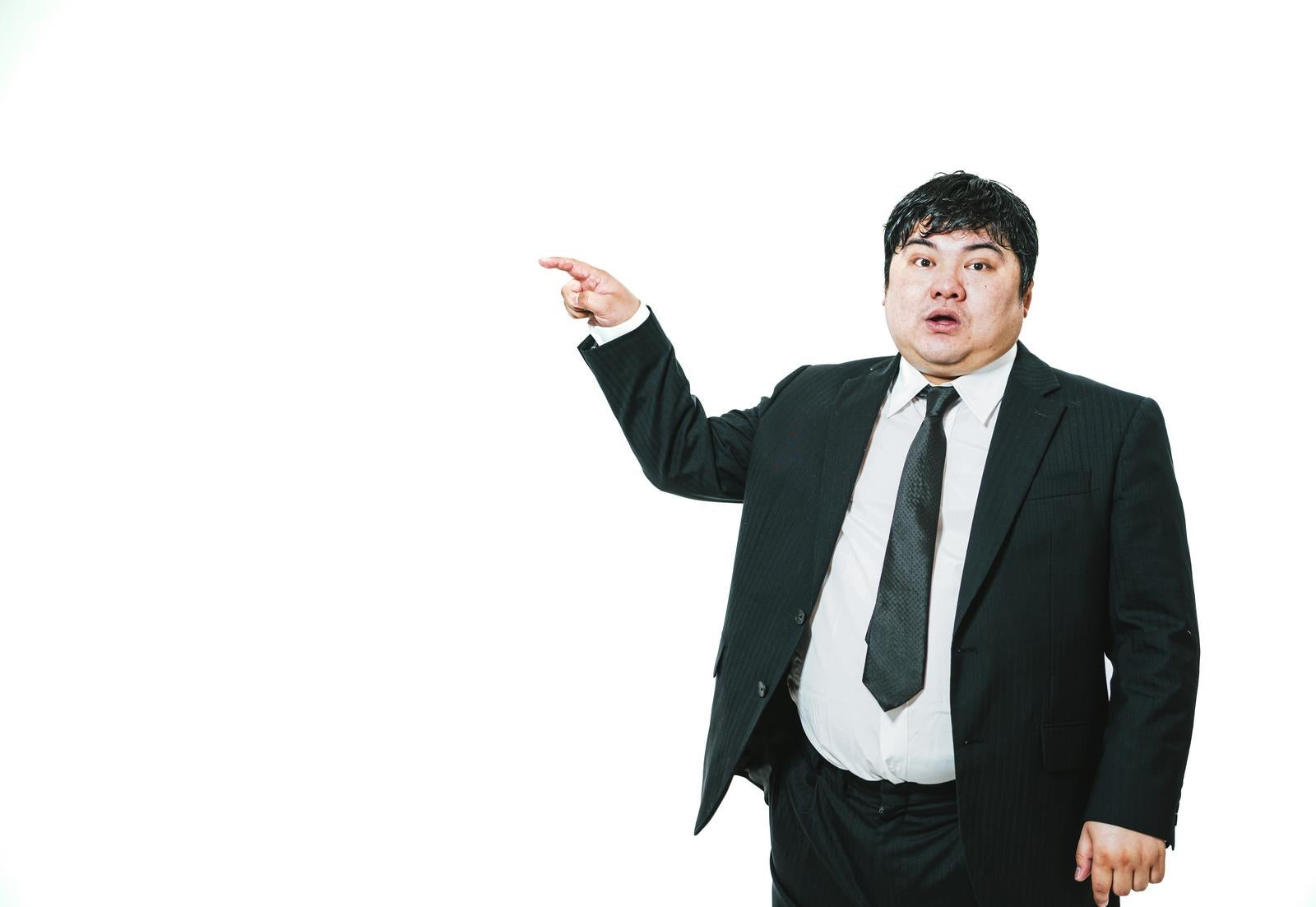 白背景で左方向を指さしながら口を開けているスーツ姿の太った男性［モデル：段田隼人］