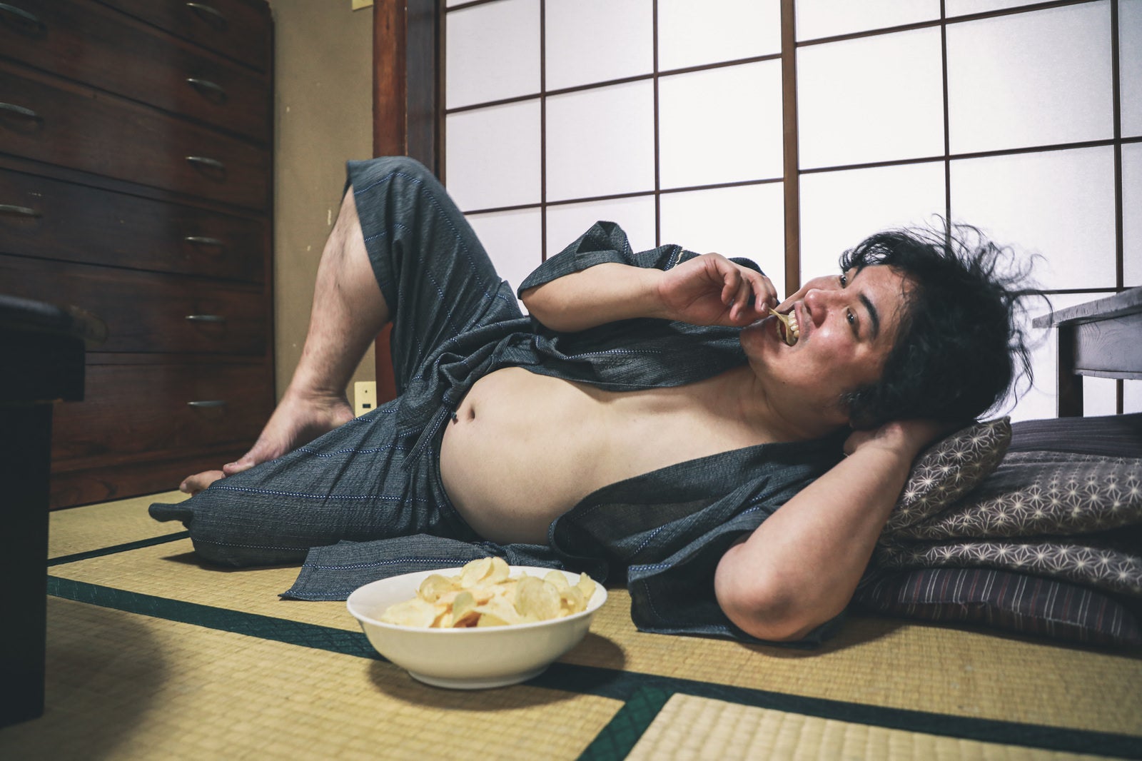 「腹を出してポテチを食べながら寝転ぶ昭和男子」の写真［モデル：段田隼人］