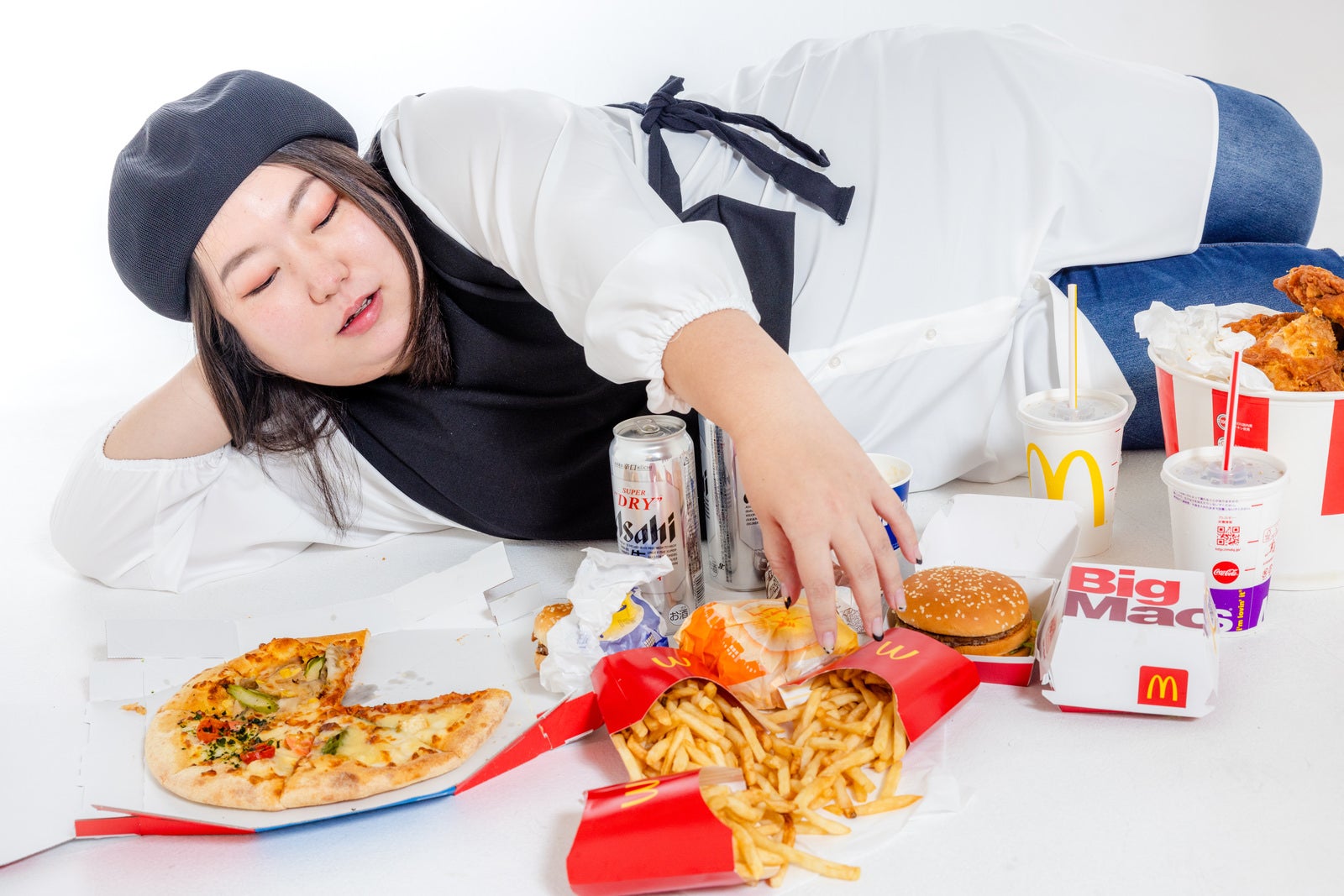 ファーストフードに囲まれテーブルの上に横たわり食べ散らかす女性［モデル：KANA］