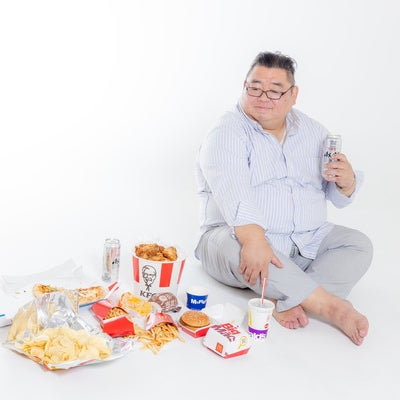 揚げ物やハンバーガーなどに囲まれてほっこりする肥満男性の写真
