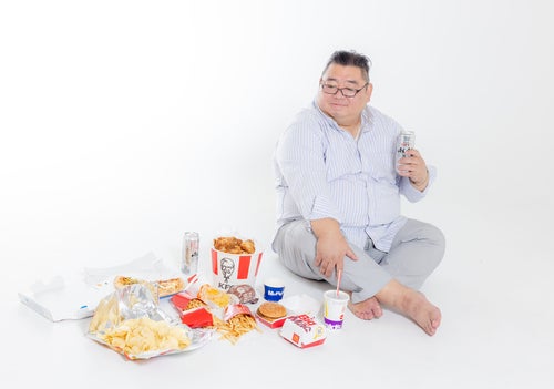 揚げ物やハンバーガーなどに囲まれてほっこりする肥満男性の写真