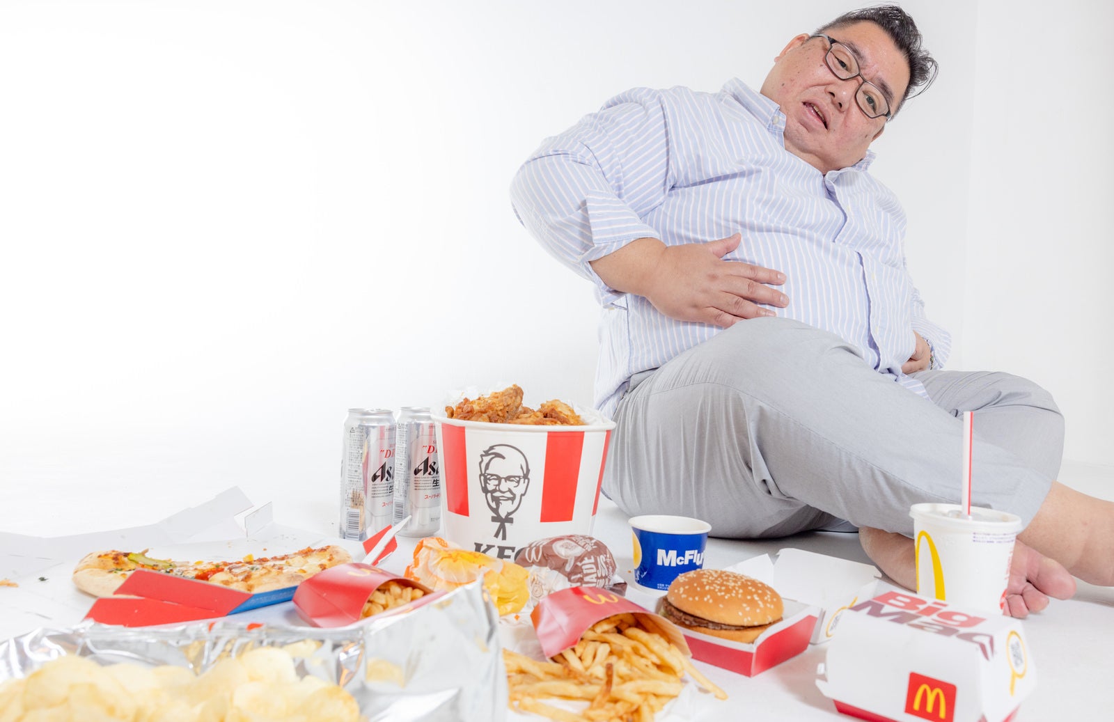 フライドチキンやピザなどのジャンクフードに囲まれて食べ過ぎでお腹を押さえる太った男性［モデル：ダルマ親方］
