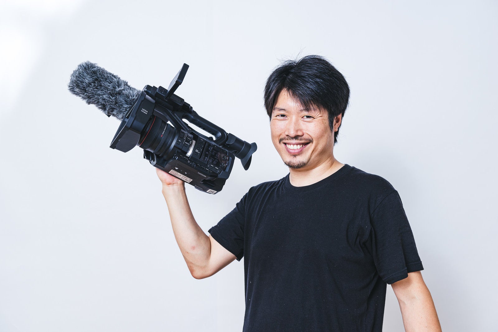 「業務用ビデオカメラを片手で持ち上げる笑顔の男性カメラマン」の写真［モデル：瀧野恵太］