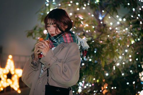 彼の分の温かい飲み物で手を温めながらクリスマスツリーの前で待つ彼女の写真