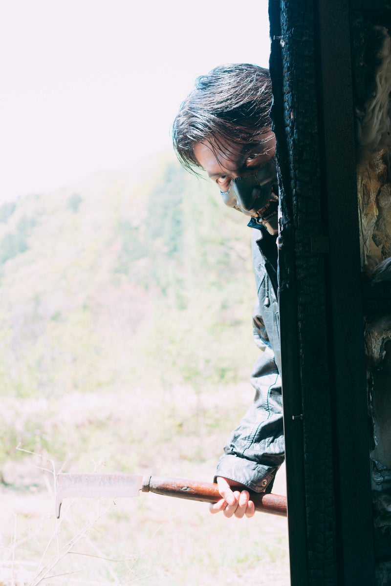 山道の古い建物の陰から周囲を伺う長髪の男性の警戒の写真