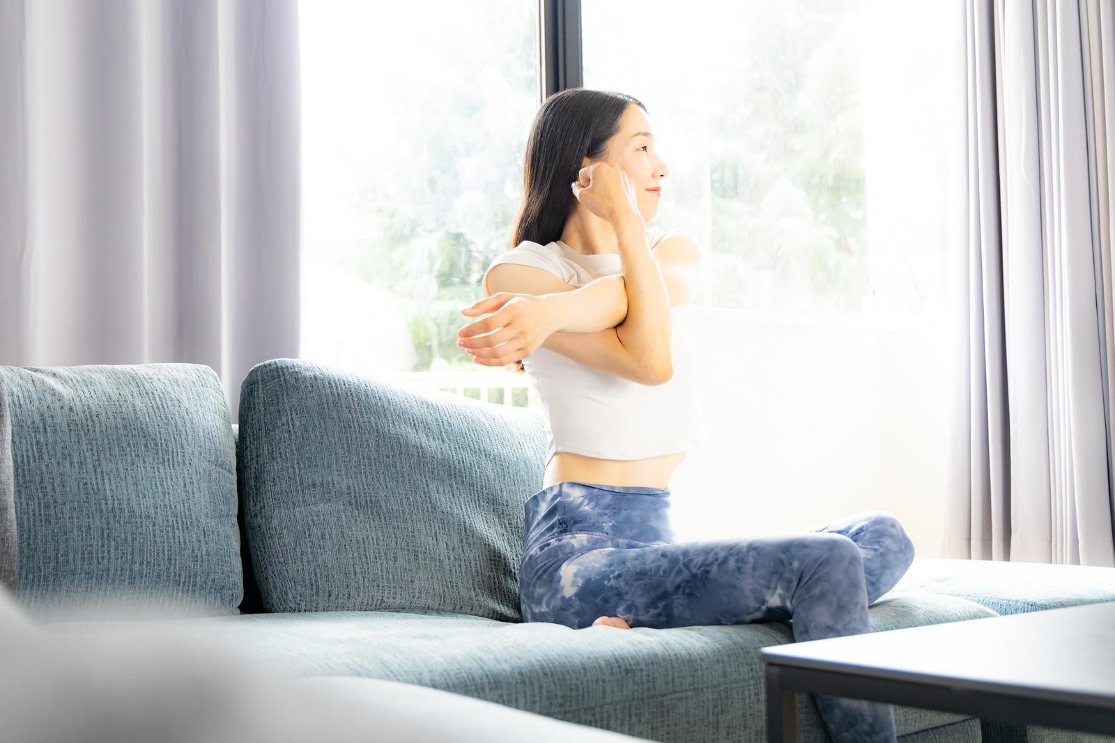 リビングのソファでストレッチ運動をする白いトップス姿の女性［モデル：ひるかわえみこ］